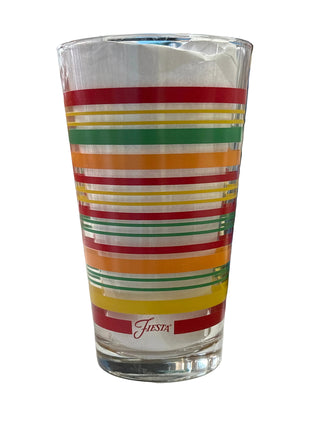 A clear glass pint glass with horizontal multicolor stripes in red, green, yellow, and orange, featuring the 'Fiesta' brand logo printed in red near the bottom. The glass is photographed on a tabletop with a blurred background containing other items.